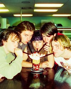 four young men sitting at a table with a cake in front of them and one man leaning on his head