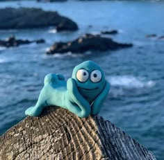 a blue stuffed animal sitting on top of a wooden log next to the ocean with rocks in the background