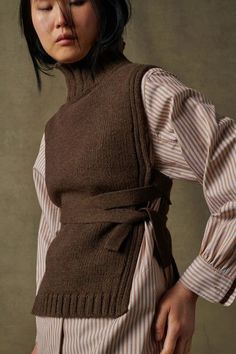 a woman with her eyes closed wearing a brown sweater and striped dress, standing in front of a gray background