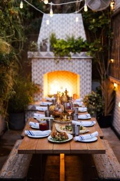 an outdoor dining table set with plates and food on it, in front of a fire place