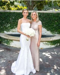 two beautiful women standing next to each other in front of a fountain with greenery