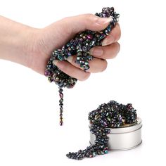 a hand holding a tin with beads in it next to a can filled with black and multicolored beads