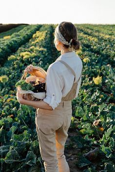 a woman standing in a field holding a basket full of vegetables and looking at the horizon