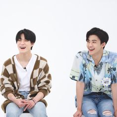 two young men sitting next to each other on top of a white surface and laughing