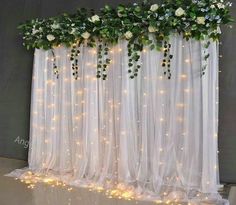 white flowers and greenery are hanging on the sheer curtain with lights in front of them