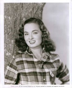 an old black and white photo of a woman in plaid shirt posing next to a tree