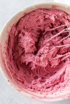 a white bowl filled with pink frosting next to a whisk in it