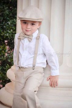 a young boy in a white shirt and bow tie standing next to a pillar wearing suspenders and a hat