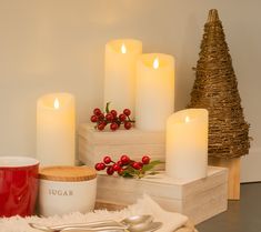 white candles are sitting on a table next to a red mug and silverware in front of a small christmas tree