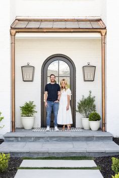 a man and woman standing in front of a door with potted plants on the steps