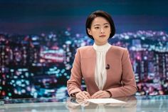 a woman sitting at a desk in front of a tv screen with the city lights behind her