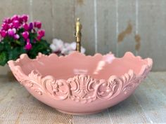 a pink bowl sitting on top of a table next to purple flowers and a gold faucet