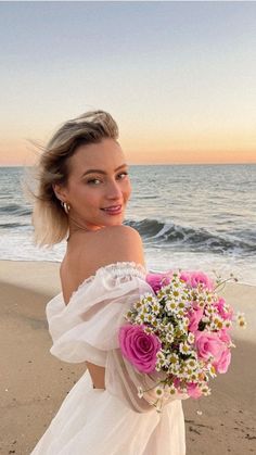 a woman in a white dress holding a bouquet of pink and white flowers on the beach