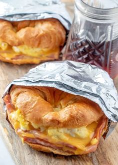 two breakfast sandwiches wrapped in aluminum foil on a cutting board next to a jar of jam