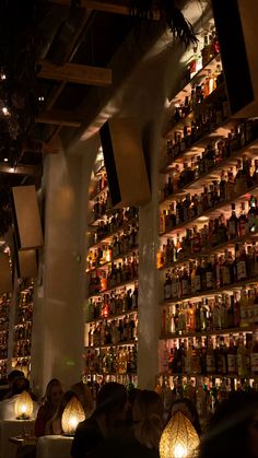 people are sitting at tables in front of shelves with bottles on them and lamps hanging from the ceiling