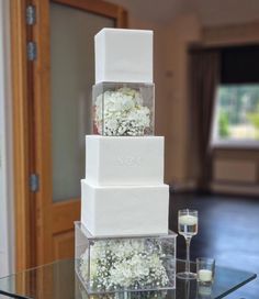 a three tiered white wedding cake with flowers on the top and bottom, sitting on a glass table in front of a doorway