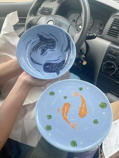 two plates with goldfish painted on them in the front seat of a car, one being held up by someone's hand