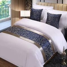 a white bed with blue and gold comforter in a bedroom next to a window