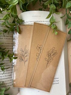 three bookmarks sitting on top of an open book next to some plants and leaves