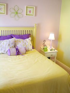 a yellow bed with purple sheets and pillows in a pink walled room, next to a white night stand