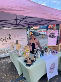 a woman standing behind a table with various items on it under a pink tent at an outdoor event