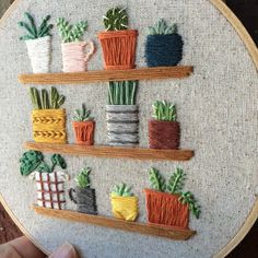 a hand embroidered wall hanging with potted plants and pots on the shelves in front of it