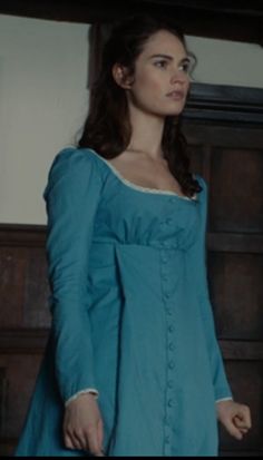 a woman in a blue dress standing next to a wooden dresser and looking off into the distance