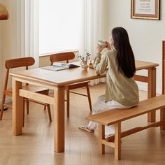 a woman sitting at a wooden table drinking from a cup and looking out the window