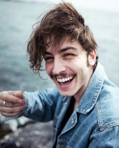 a man with a smile on his face sitting by the water and holding something in his hand