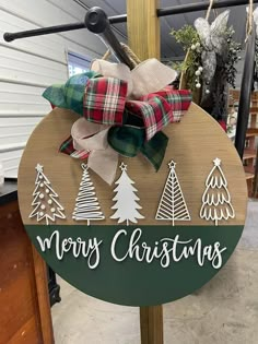 a merry christmas sign with plaid bows and evergreen trees on the front, hanging from a wooden pole