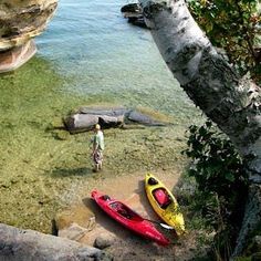 two kayaks are sitting in the water next to some rocks and trees, while a man stands near them