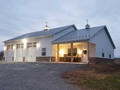 a large white barn with lights on it's doors and windows in the evening