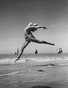 a woman jumping into the air while standing on top of a beach next to the ocean