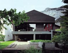 the house is surrounded by lush green trees and mountains in the background, with an outdoor dining area on the upper level