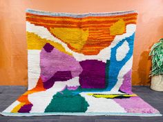 a multicolored rug on the floor next to a potted plant in front of an orange wall