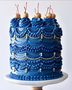 a blue cake with gold decorations and cherries on top is sitting on a pedestal