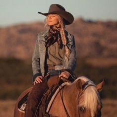 a woman riding on the back of a brown horse in a field with mountains in the background