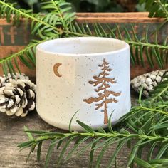 a white and gold mug with pine trees on it sitting next to pine cones, evergreen branches