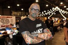 a man with tattoos and piercings standing in a room full of other tattooed people