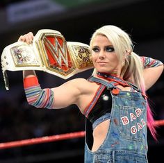 a woman in overalls holding up a wrestling belt and looking at the camera with her arm raised