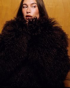 a woman with long hair wearing a black fur coat and posing for the camera in front of a wooden wall