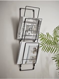 two metal racks holding magazines and papers on a wall next to a plant in a vase