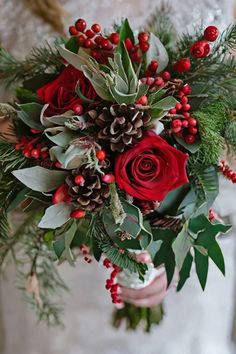 a bride holding a bouquet of red roses and greenery with pine cones on it