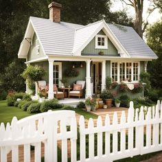 a white picket fence surrounding a small green house with flowers in the window boxes and potted plants on the front porch