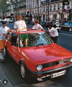 three people standing next to a red car on the side of a road with buildings in the background