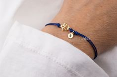 a person wearing a blue cord bracelet with two gold charms on the clasp and an initial charm