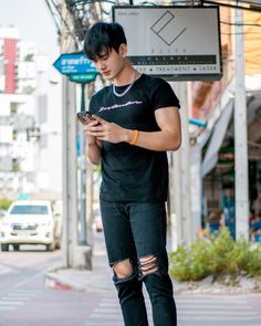 a young man standing on the street looking at his cell phone while wearing ripped jeans