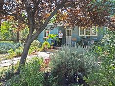 a man standing in the doorway of a blue house surrounded by trees and plants on a sunny day