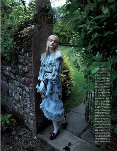 a woman standing in front of a brick wall wearing a blue dress and black shoes