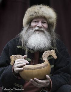 an old man with a long beard holding a wooden object in his hands and wearing a fur hat